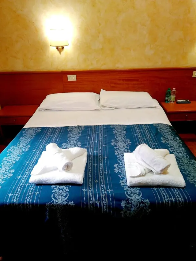 A double bed with a blue patterned spread and two folded towel stacks sits beside a wall lamp and bedside tables holding water bottles.
