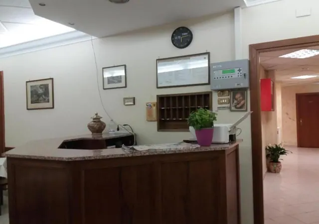 A wooden reception desk with a granite top stands in a lobby, with a wall clock, framed pictures, a pigeonhole cabinet and a potted plant on the counter.