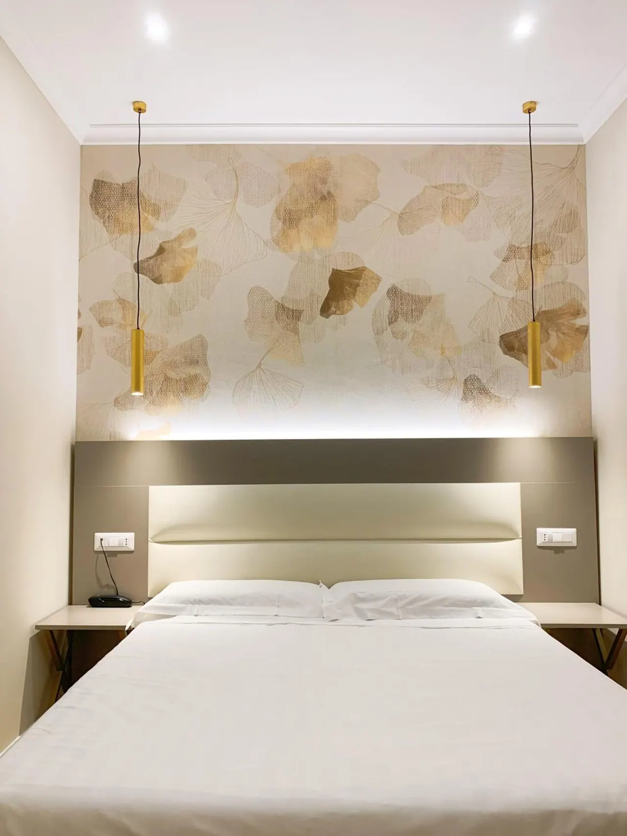 A bedroom with a double bed under white linen, a headboard wall with leaf-patterned wallpaper, and two gold pendant lights hanging from the ceiling.