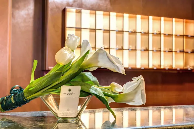 White tulips and calla lilies sit in a glass vase on a marble reception counter, with a hotel card tucked into the arrangement and illuminated shelving behind.