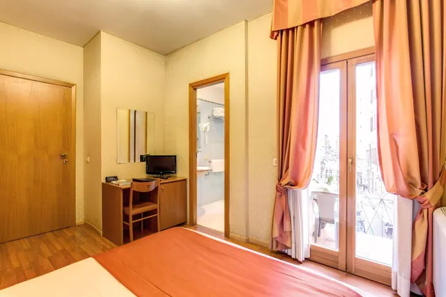 A hotel bedroom with a wooden desk and television, a doorway to the bathroom, and French doors opening onto a balcony with white chairs and neighbouring buildings beyond.