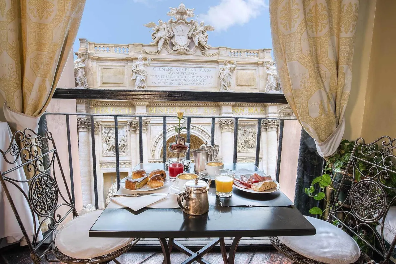 A breakfast table with pastries, orange juice and a silver coffee pot sits on a balcony overlooking the Trevi Fountain facade.