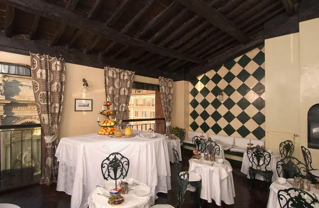 A dining room with white-clothed round tables, wrought iron chairs, a green and cream checkered wall and a beamed wooden ceiling, with a fruit stand on one table.