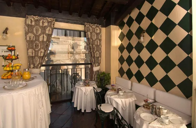 A breakfast room with white-clothed tables, a checkered green and cream wall, patterned curtains and a window onto a classical building facade.