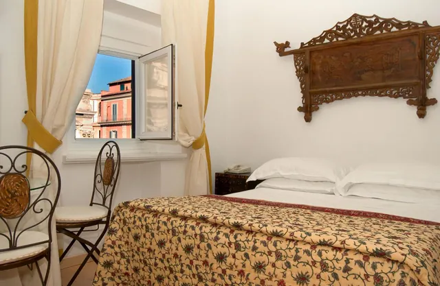 A bedroom with a double bed under a carved wooden headboard, with an open window beside it showing pink and ochre buildings outside.