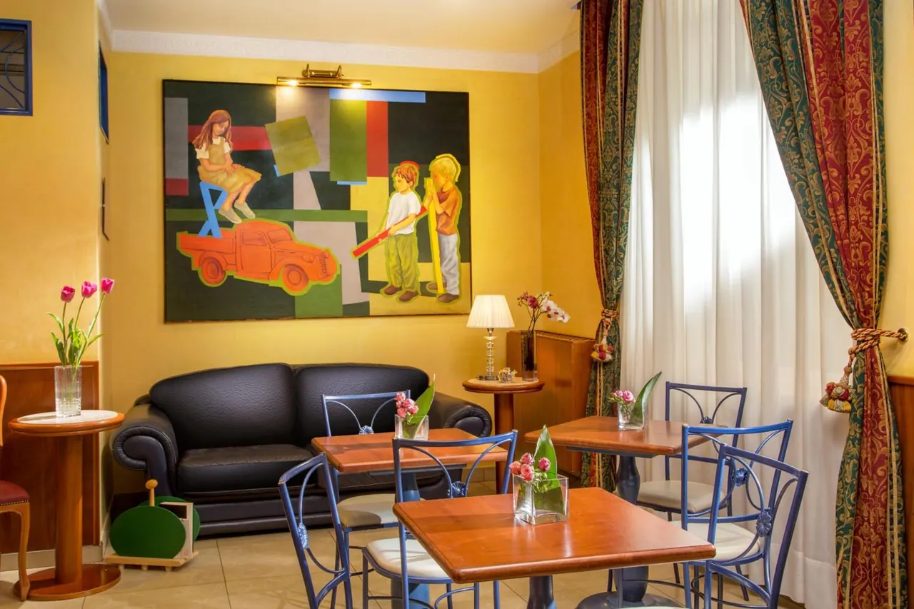A seating area with a black leather sofa, a large painting on a yellow wall, and small wooden tables with blue metal chairs beside sheer white curtains.