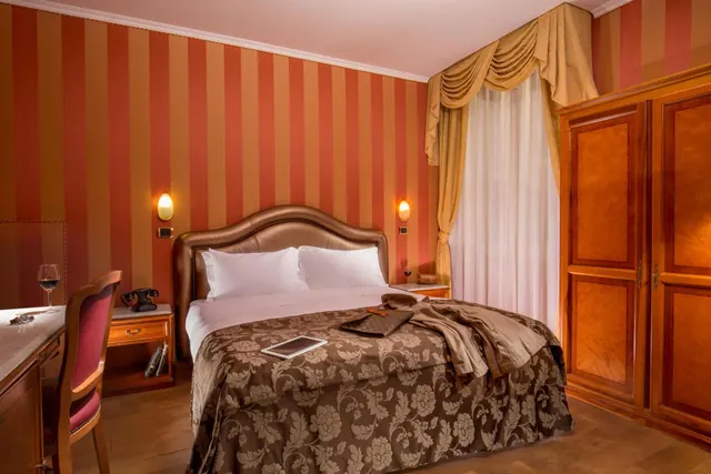 A double bed with a brown leather headboard and floral throw sits beside a wooden wardrobe, a desk with a wine glass, and a telephone on a nightstand in a room with striped wallpaper.