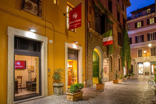 Evening street view of the Hotel Forte facade with illuminated entrances, potted plants, a red hotel flag and an Italian flag on a cobbled street.