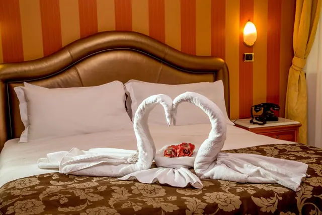 A made-up double bed with a brown leather headboard, white towel swans forming a heart with red flowers, and a black telephone on a wooden nightstand beside striped orange wallpaper.