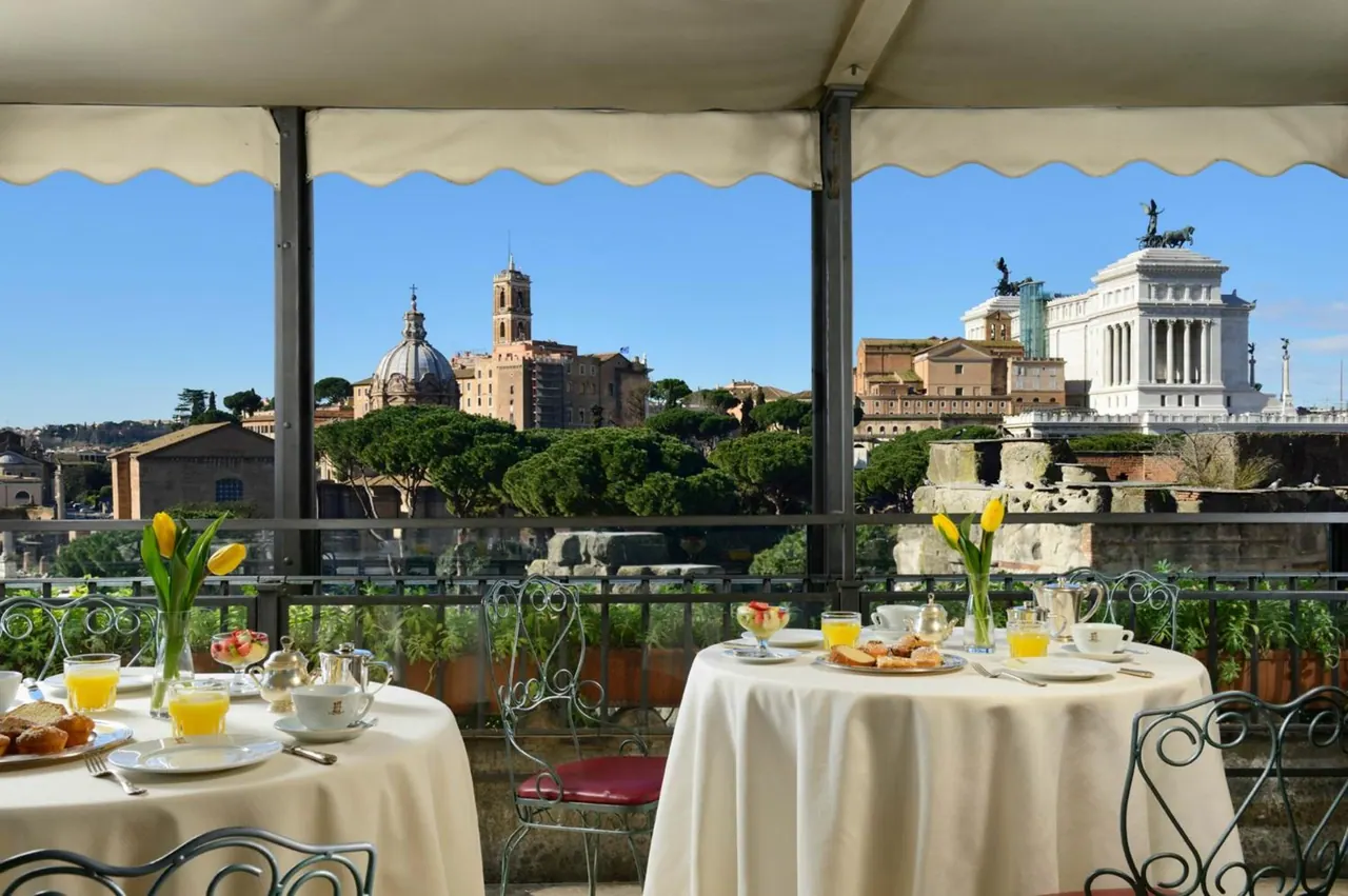 Two breakfast tables with white linen and wrought iron chairs sit under an awning on a terrace overlooking domes, ancient ruins and the white Vittoriano monument.