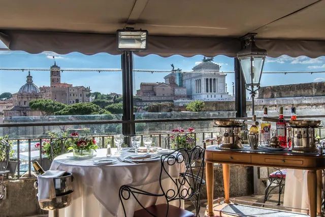 A set dining table on a covered rooftop terrace overlooks the white Vittoriano monument and domed churches across Rome.