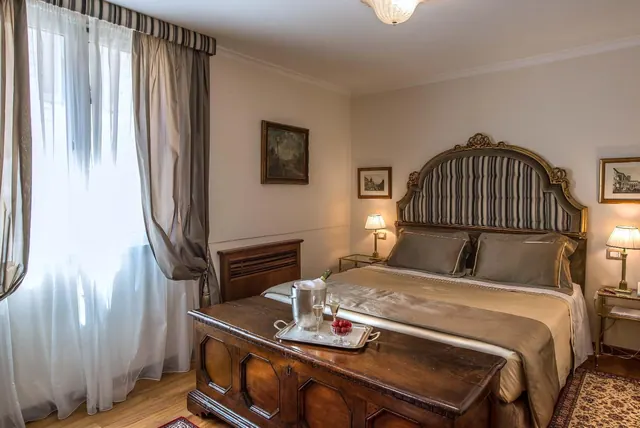 A bedroom with a double bed under a striped upholstered headboard, a wooden chest at the foot of the bed holding a tray with a champagne bucket, glasses and a bowl of fruit, and a window with sheer curtains.