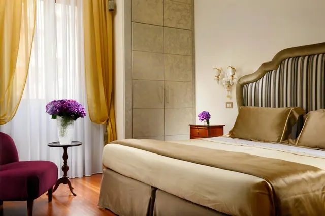 A bedroom with a large bed with a striped upholstered headboard, a magenta armchair, a small round side table with purple flowers, and a tall wall cabinet beside sheer white and gold curtains.