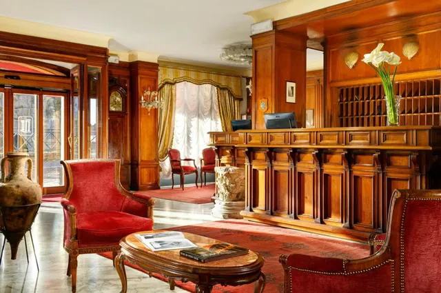 A hotel lobby with dark wood-panelled reception desk, red velvet armchairs, a marble-topped coffee table, a grandfather clock and a vase of white flowers.