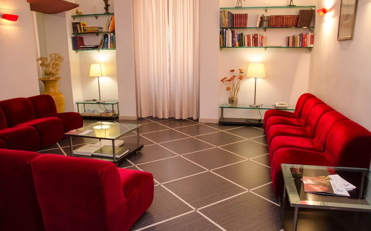 A lobby seating area with red velvet sofas, glass coffee tables, bookshelves, table lamps and a curtained window.