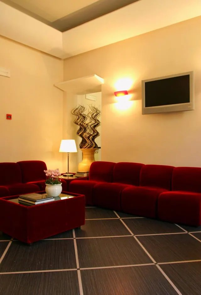 A seating area with red velvet modular sofas, a red coffee table holding books and an orchid, a table lamp, and a wall-mounted flat-screen television that is switched off.
