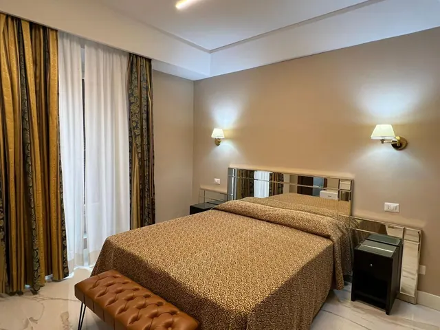 A double bed with a gold patterned bedspread sits beside a mirrored headboard, with wall sconces above and gold striped curtains by the window.