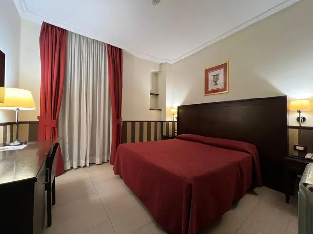 A bedroom with a double bed under a red bedspread, red and white curtains, wall lamps, and a desk with a chair on the left.