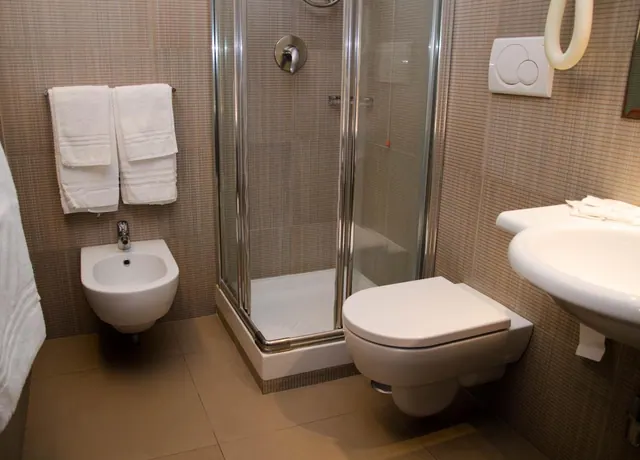 A bathroom with a glass-enclosed corner shower, a wall-hung toilet and bidet, a wall-mounted sink, and white towels hanging on a rail.