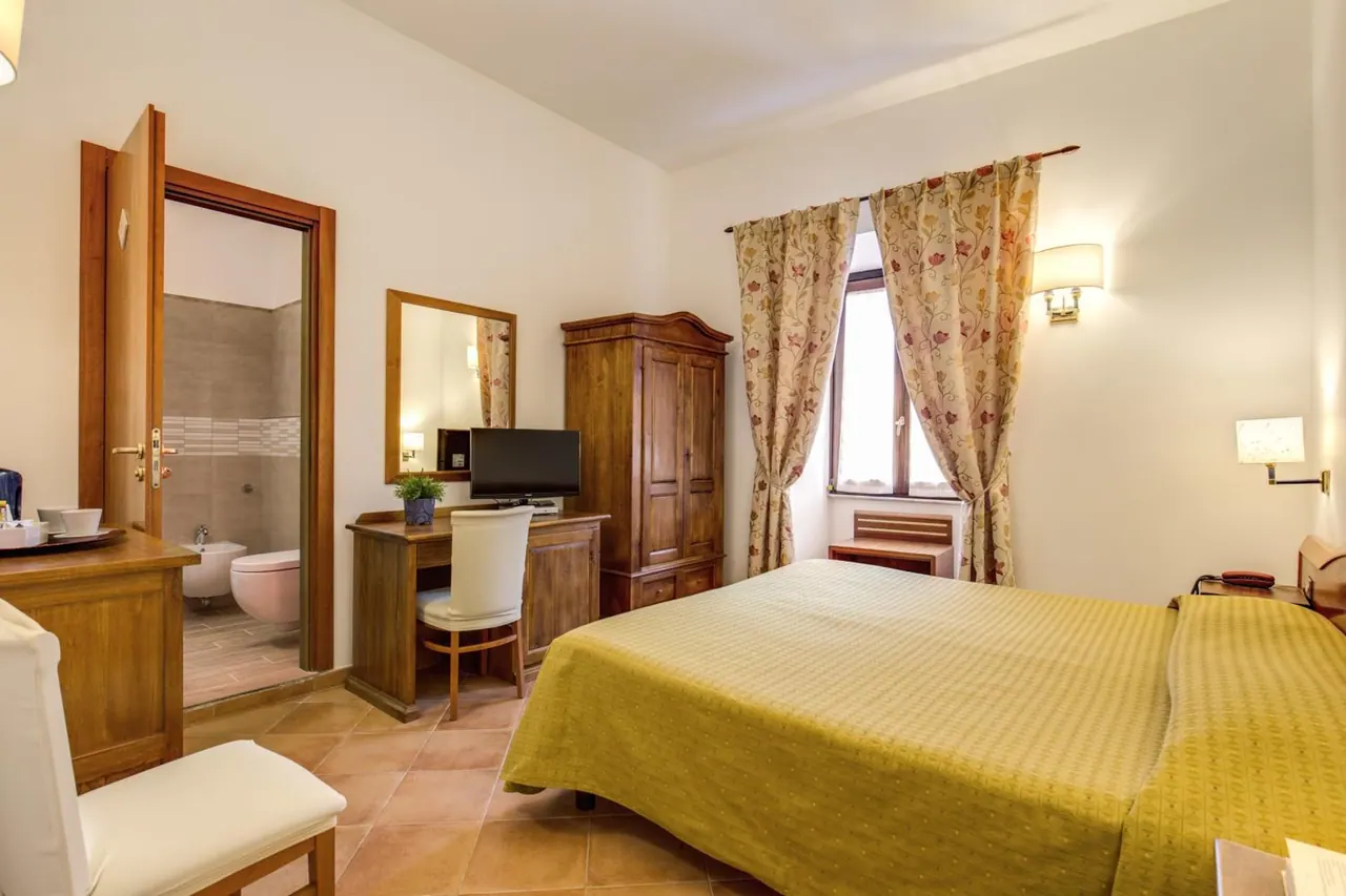 A double bed with a yellow bedspread sits beside a wooden wardrobe and desk with a television, and an open door shows an en-suite bathroom with a bidet and toilet.
