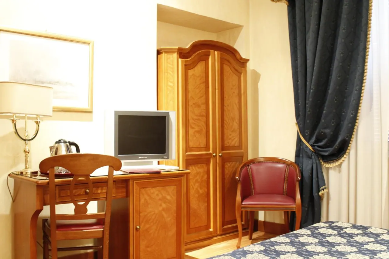 A hotel room with a wooden desk and chair, a table lamp, a small CRT television, an electric kettle, a wooden wardrobe, a red leather armchair and dark blue curtains.