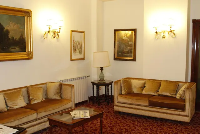 A hotel lounge seating area with two velvet sofas, framed paintings, wall sconces, a side table with a lamp, and a glass-top coffee table holding newspapers.