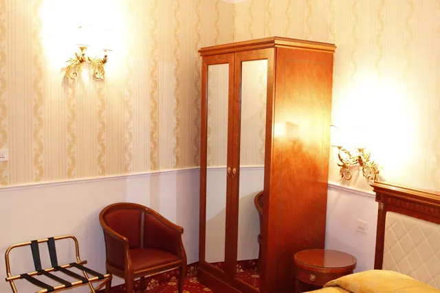 A hotel bedroom with striped wallpaper, a wooden wardrobe with mirrored doors, a red leather armchair, a luggage rack and a bed with a quilted white headboard at the right edge.