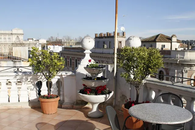 A rooftop terrace with a white balustrade, potted trees, a tiered stone planter and a round granite table with chairs, overlooking neighbouring rooftops and buildings under a clear sky.