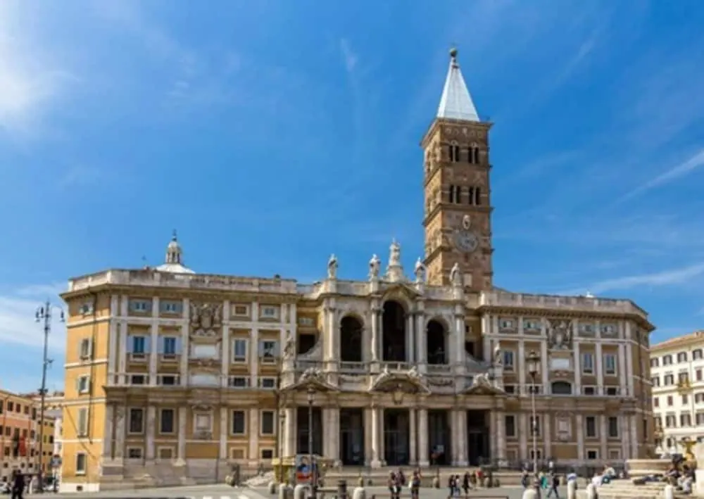 The facade and bell tower of the Basilica di Santa Maria Maggiore stand under a clear blue sky, with a few people walking in the piazza below.