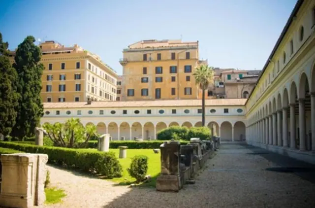 A courtyard with a long arcaded portico, a gravel path, a lawn, clipped hedges, stone fragments and ochre apartment buildings behind.
