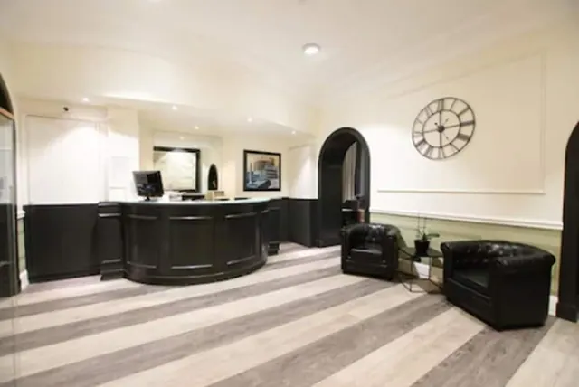 A curved black reception desk with a computer monitor stands in a lobby beside two black leather armchairs, a small glass side table with a plant, and a large wall clock.