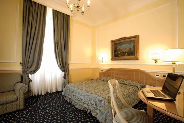 A double bed with a blue patterned cover sits beside a tall window with grey curtains, an armchair, a wooden desk with a laptop, and a framed painting on the wall.