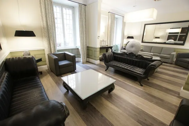 A lounge seating area with black leather sofas, an armchair, a glass-topped coffee table, a large wall mirror and tall curtained windows.