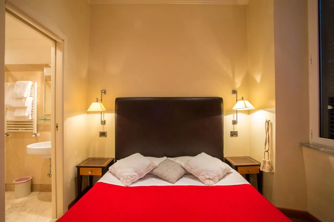 A double bed with a red throw and decorative pillows sits in a warmly lit room, with the bathroom visible through an open door on the left.