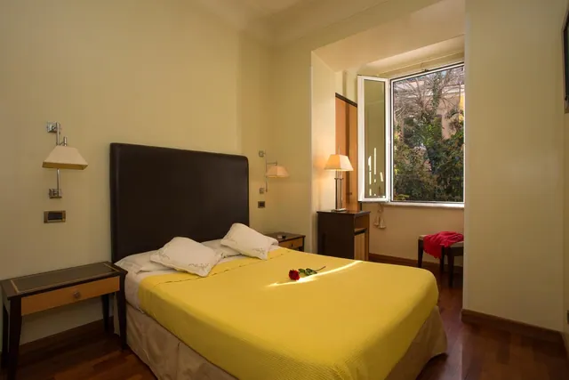 A double bed with a yellow bedspread and a red rose on it, with bedside lamps and an open window showing trees outside.