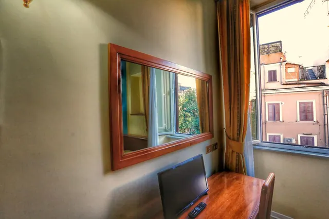 A wooden desk with a flat-screen television and remote control sits beside a window that looks onto a pink building across the street, with a framed mirror on the wall.