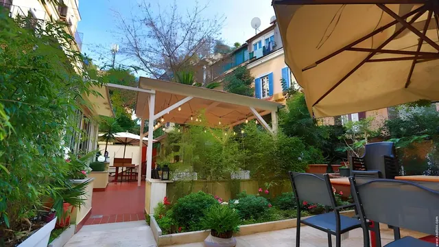 A garden courtyard with a covered pergola strung with lights, shade umbrellas, outdoor chairs and tables, and lush potted plants.