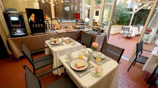 A breakfast room with set tables, a pastry buffet, a coffee machine and juice dispensers, with glass doors opening onto a courtyard garden.