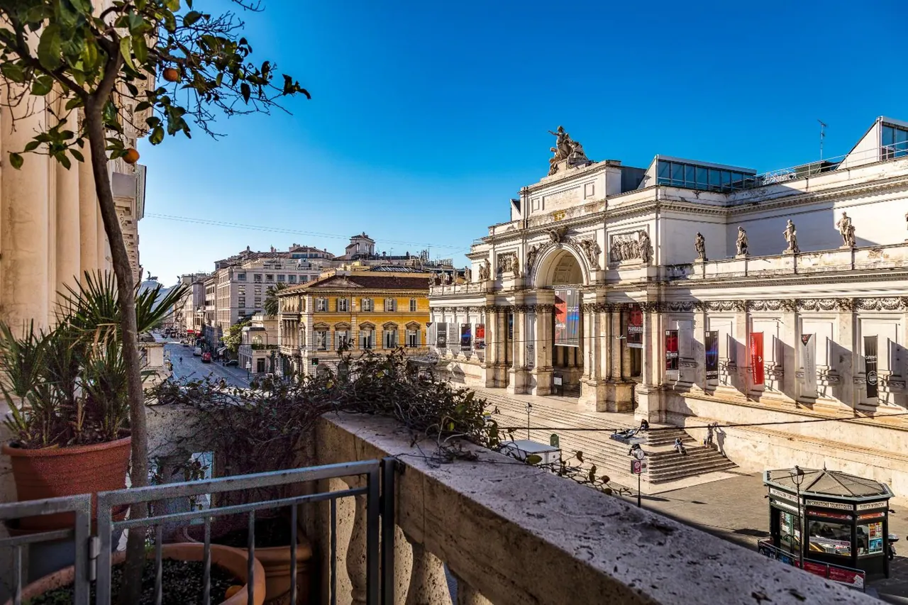 View from a balcony with a potted orange tree and travertine railing over a street toward a white neoclassical building with a sculpted chariot on its roof and a yellow building beside it.