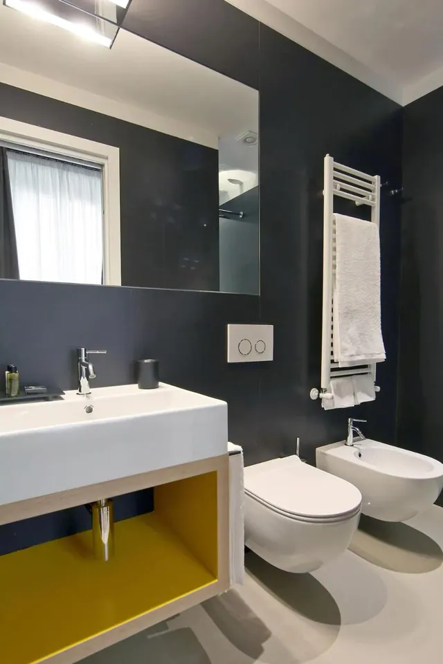 A modern bathroom with a wall-hung sink above a yellow cabinet, a wall-hung toilet and bidet, a heated towel rail, and a large mirror against navy walls.