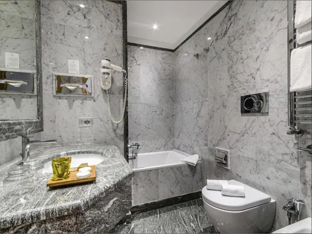 A marble-tiled bathroom with a sink on a stone counter, a built-in bathtub, a wall-hung toilet with towels on the lid, and a heated towel rail.