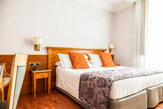A double bed with orange cushions and a floral throw sits in front of a wooden headboard, with a bedside table, wall lamps and a window with sheer curtains in the room.
