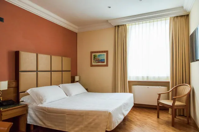 A double bed with white linen sits in a bedroom with a wooden armchair, a wall-mounted television, and a white air conditioning unit under a window with sheer curtains.