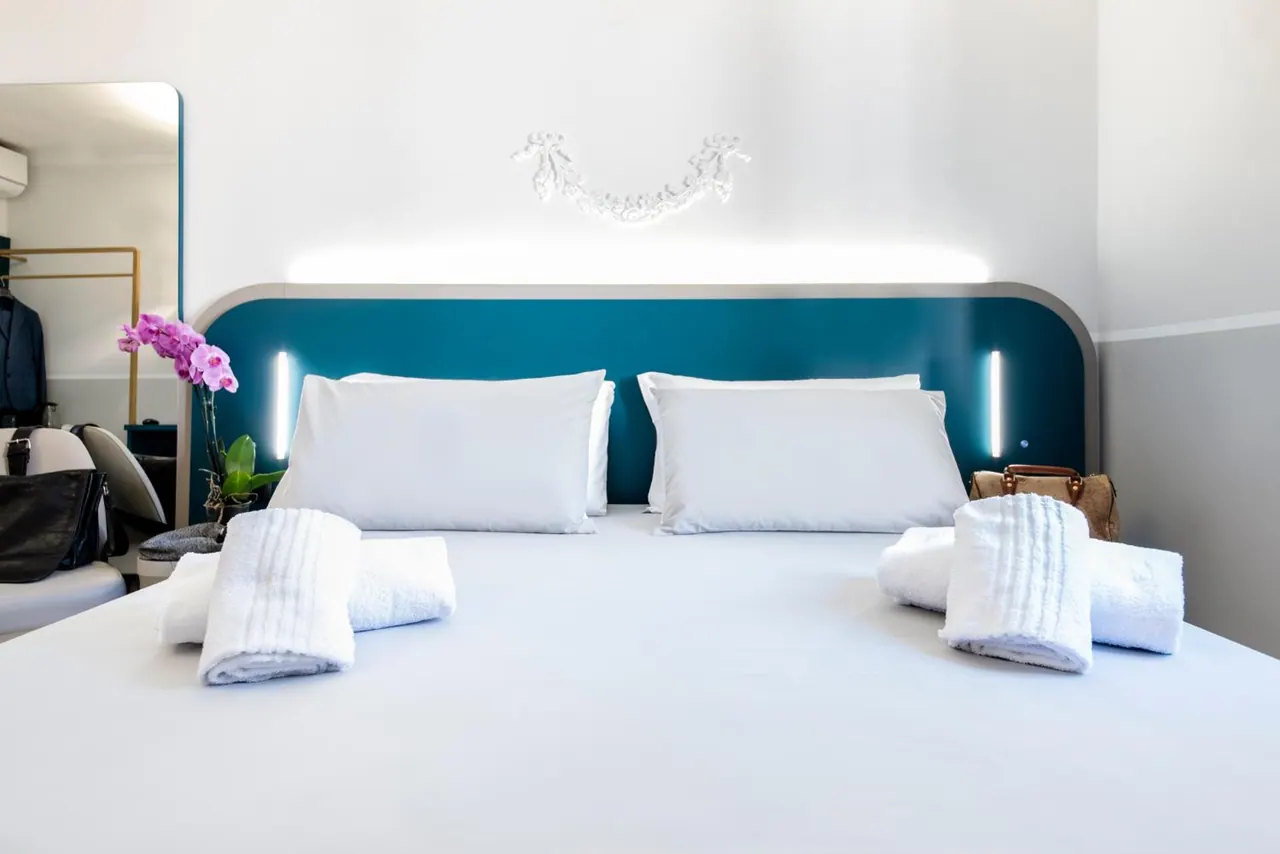 A made double bed with white linen and rolled towels sits in front of a teal headboard with wall lighting, beside a mirror and a pink orchid.