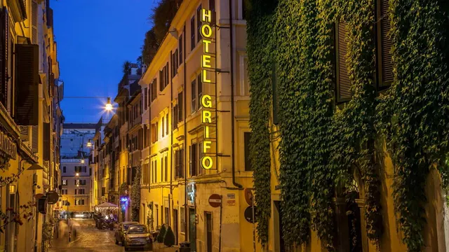 Evening view of a narrow Rome street with the vertical Hotel Grifo sign on an ivy-covered yellow building and cars parked at the curb.