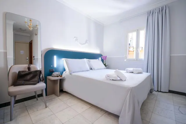 A bedroom with a double bed with white linen and a blue headboard, a tall mirror, an upholstered chair holding a bag, and a window with grey curtains.