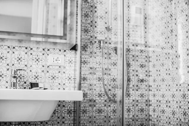 A black and white photo of a bathroom with patterned tiled walls, a wall-mounted sink, a framed mirror and a glass-enclosed shower with a handheld shower head.
