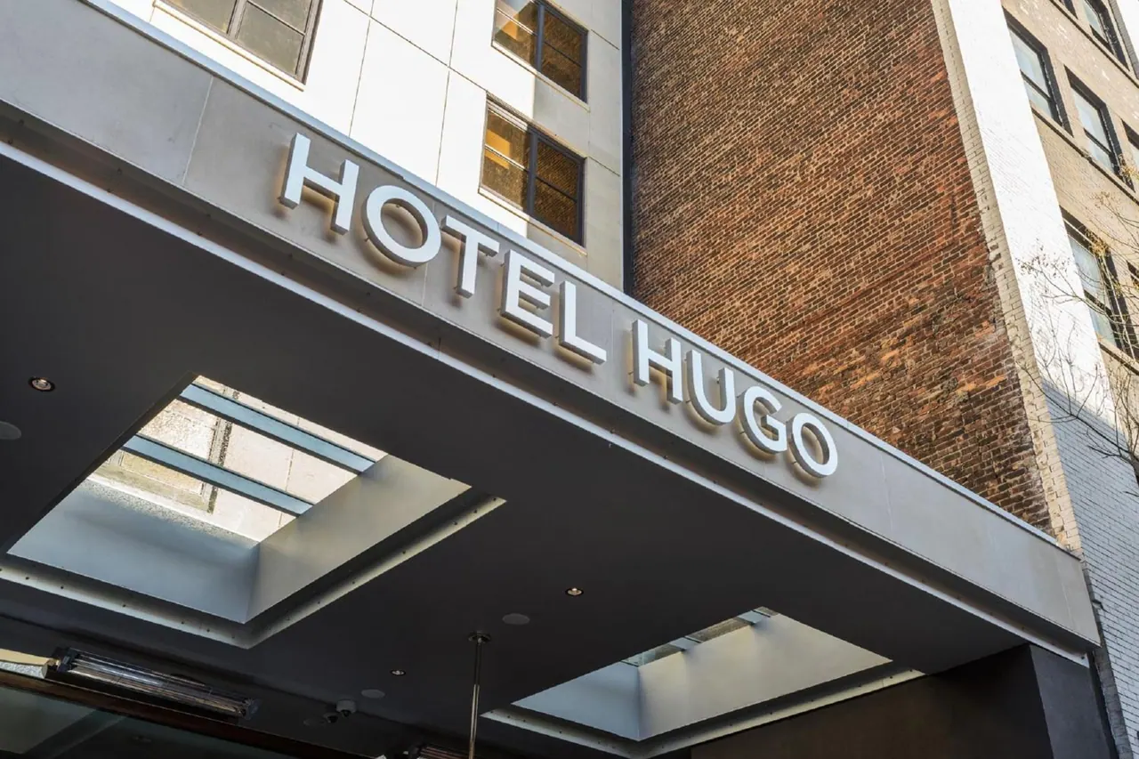 The exterior of Hotel Hugo shows its illuminated name sign on a canopy above the entrance, with a brick facade and taller building behind it.