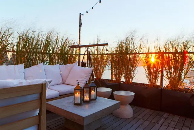 A rooftop terrace with a white sofa, a concrete coffee table holding candle lanterns, tall grasses in planters, and water visible beyond the railing at sunset.