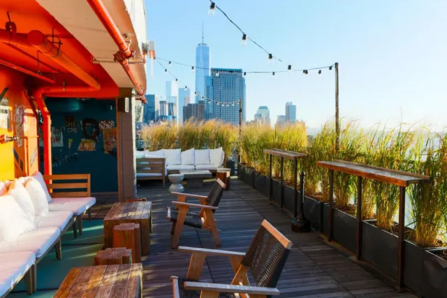 A wooden rooftop terrace with outdoor seating, planters of tall grasses and string lights, with a city skyline including One World Trade Center in the background.